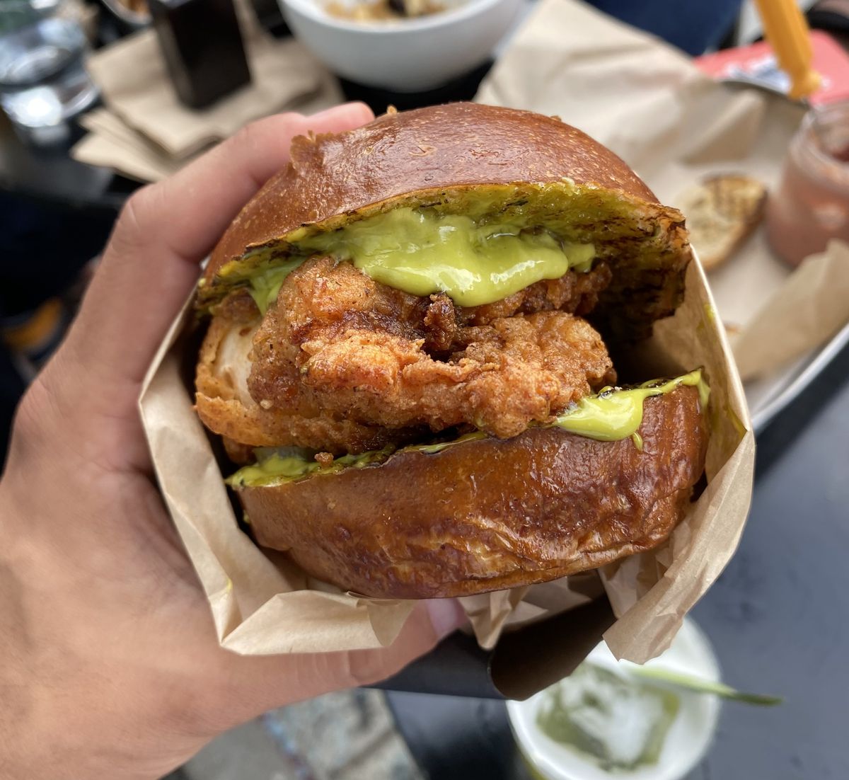 A hand holds a sandwich overflowing with green sauce and crispy chicken, in what appears to be a pretzel bun