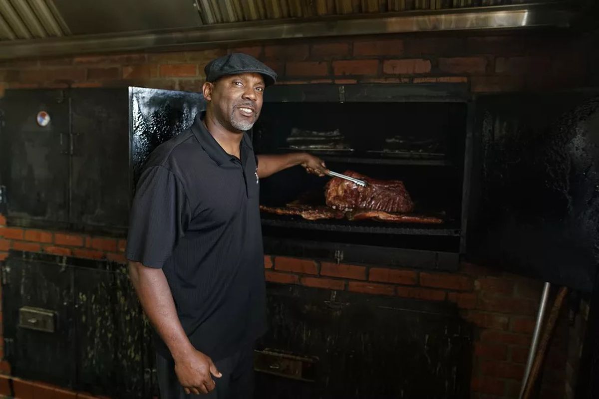 Bradrick Cooper of Coop’s West Texas BBQ standing in front of a smoker holding a rack of ribs