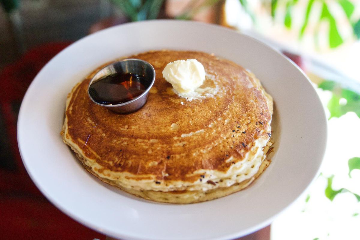 Two pancakes piled on top of each other on a white plate with a dollop of butter and a container of syrup. 