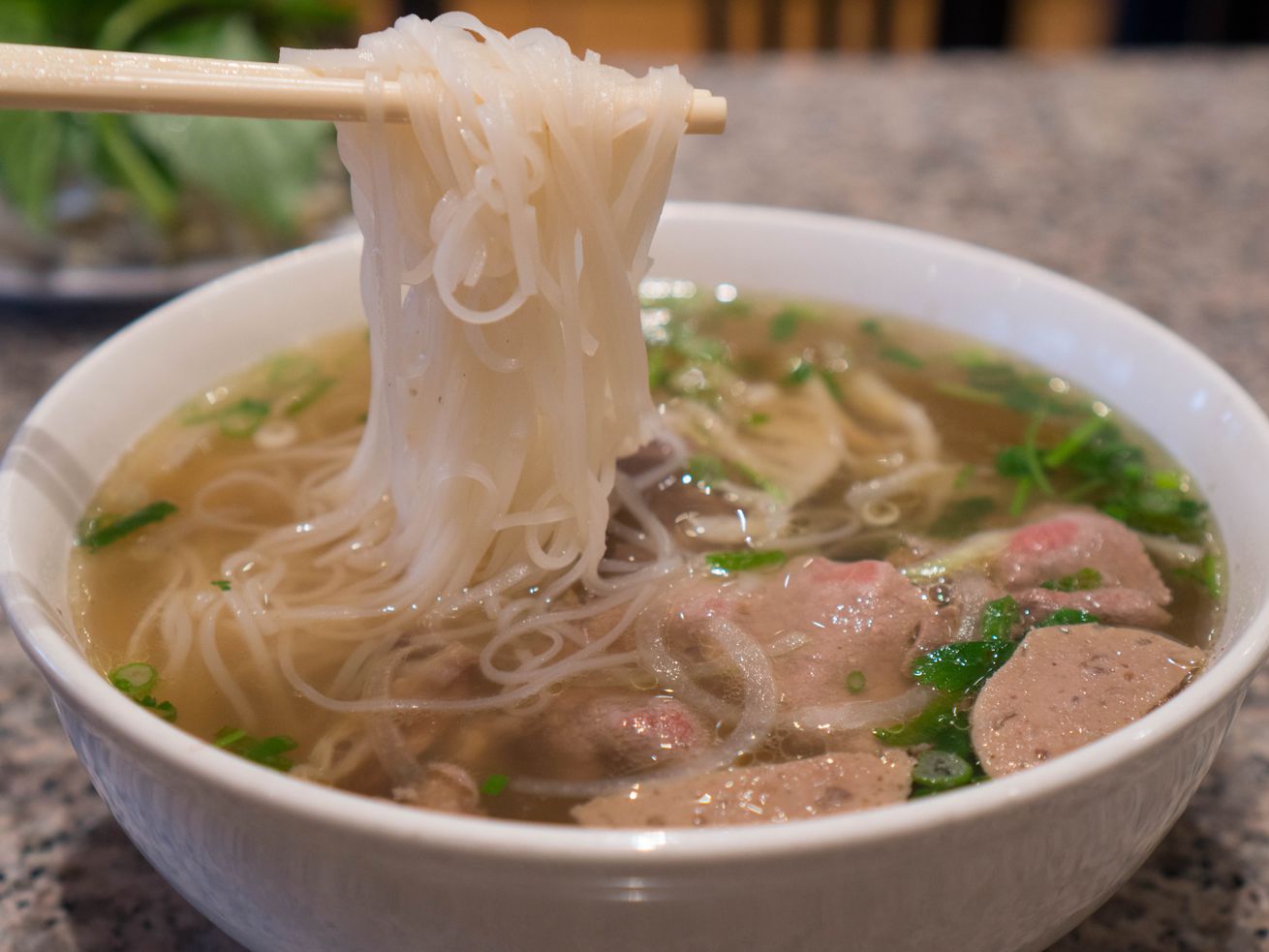 A pair of chopsticks hold up a tangle of noodles above a bowl of pho, with pieces of tripe and meatballs floating among chopped cilantro and basil