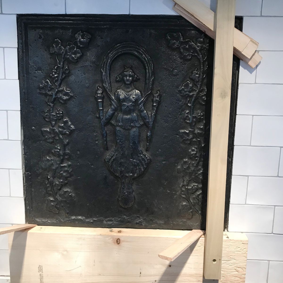 <p>The antique fireback in place as a kitchen backsplash set among white subway tile, secured temporarily with shims.</p>