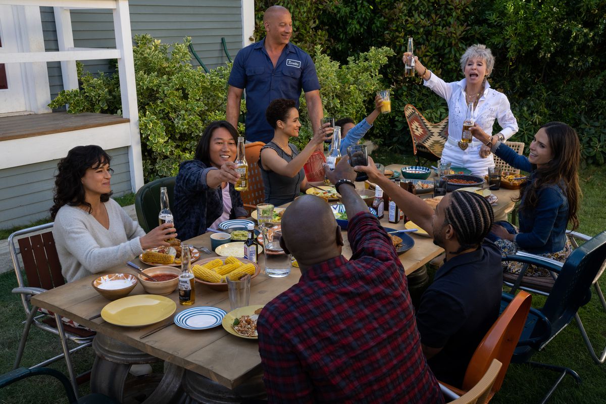 The Fast and Furious family (2023 edition) gathers around an outdoor picnic table for one of their signature cookouts in Fast X. Present and making a toast with various beers and drinks: Letty (Michelle Rodriguez), Han (Sung Kang), Ramsey (Nathalie Emmanuel), Dom (Vin Diesel), Little Brian (Leo Abelo Perry), Abuelita (Rita Moreno), Mia (Jordana Brewster), Tej (Chris ‘Ludacris’ Bridges, back to camera) and Roman (Tyrese Gibson)