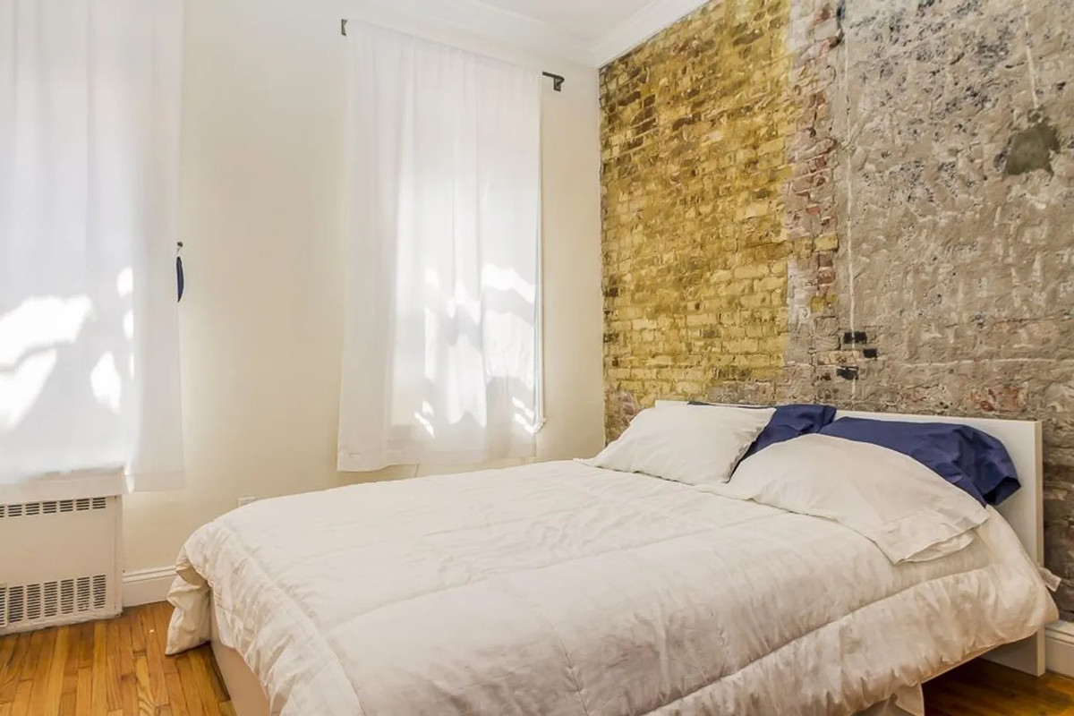 A bedroom with a large bed, exposed brick, and two large windows with white curtains.