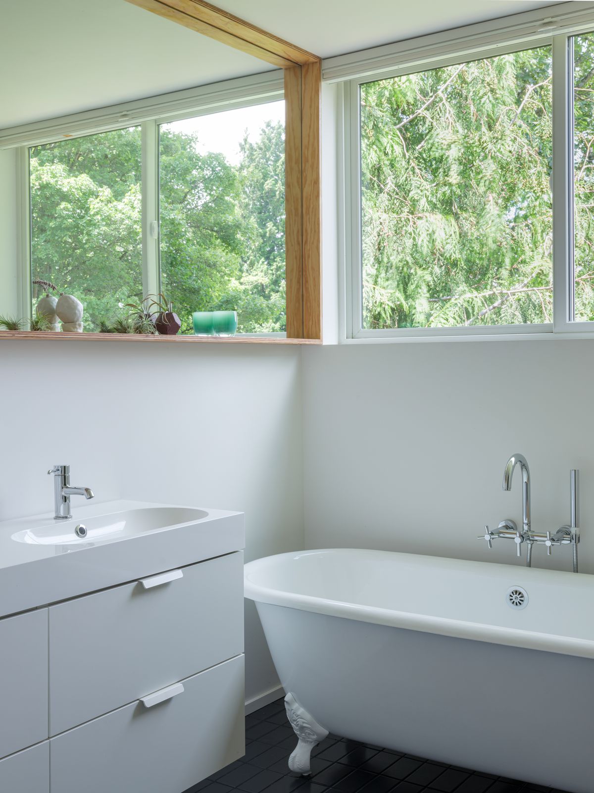 A bathroom with a bathtub, sink, cabinets, and large mirror. There are windows over the bathtub which are overlooking a view with trees.