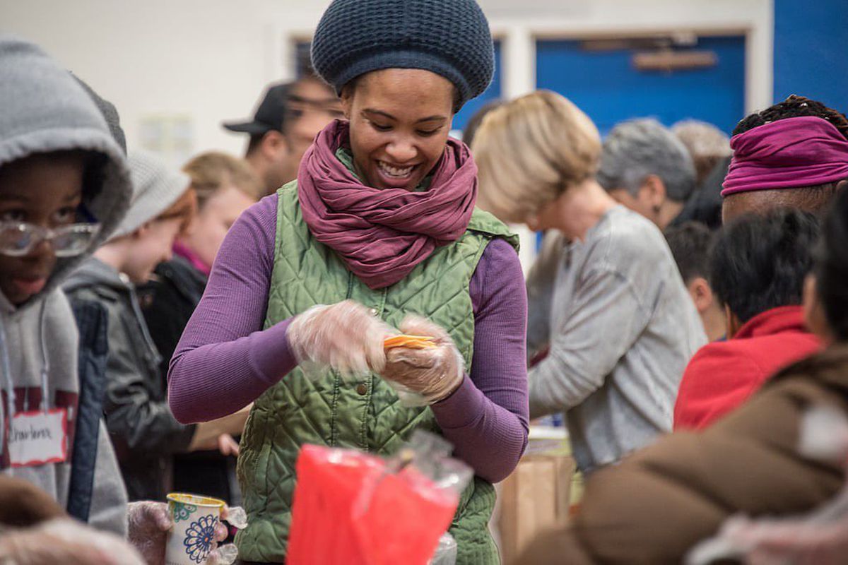 A person in a sweater and hat prepares food in a room full of people.