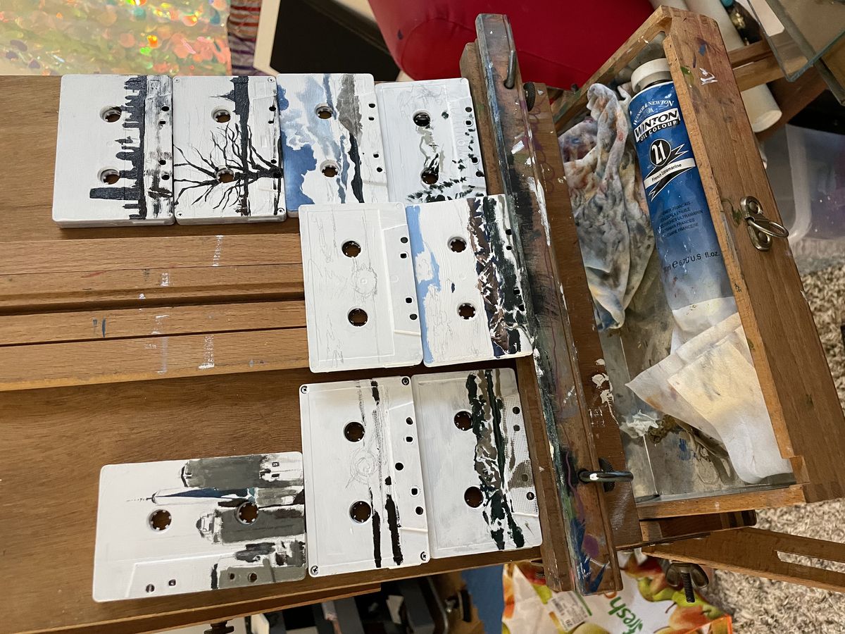 A photograph of an easel with several cassettes in the process of being painted with landscape imagery