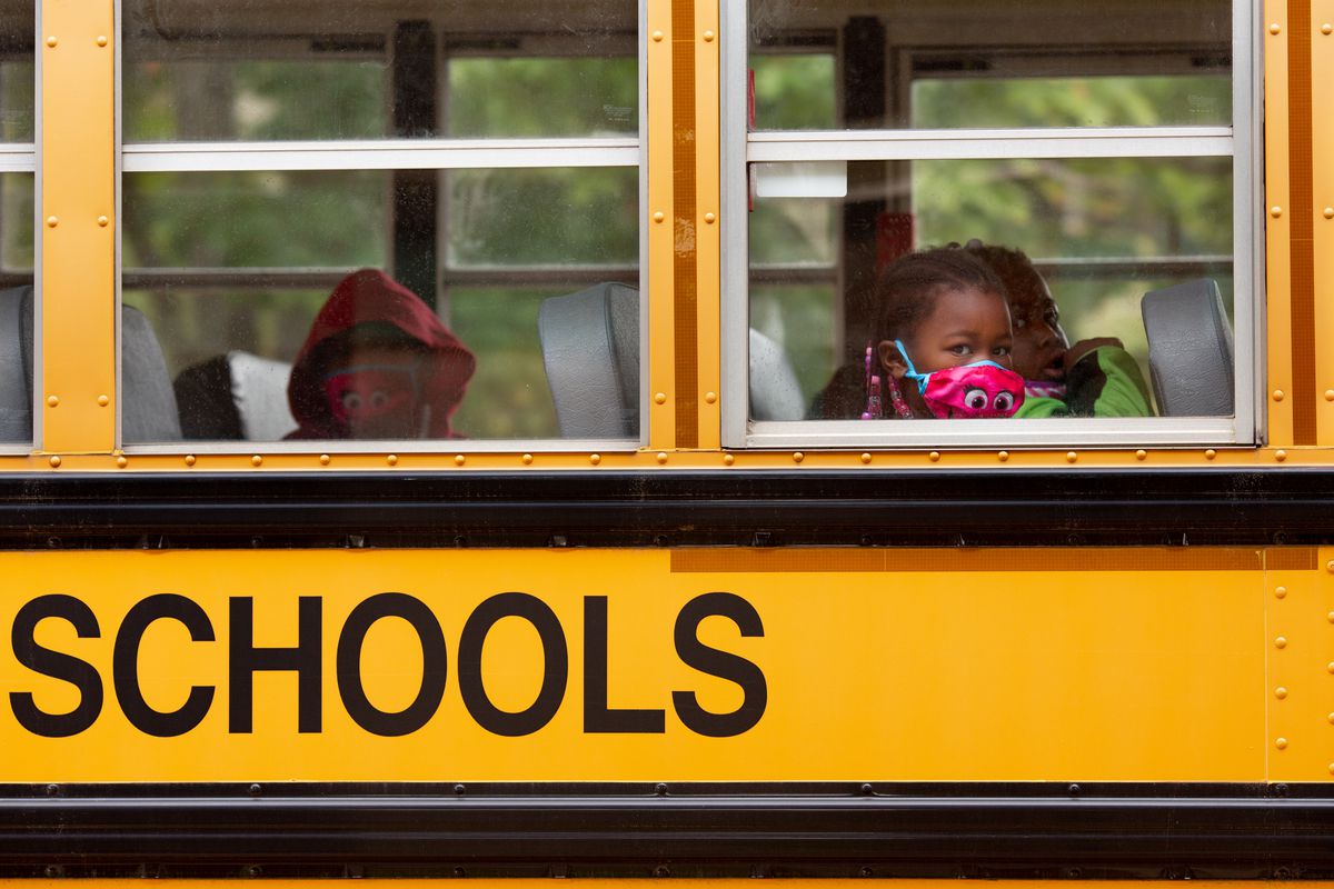 Close-up from outside a yellow school bus shows three young students wearing colorful face  masks sitting in two rows.