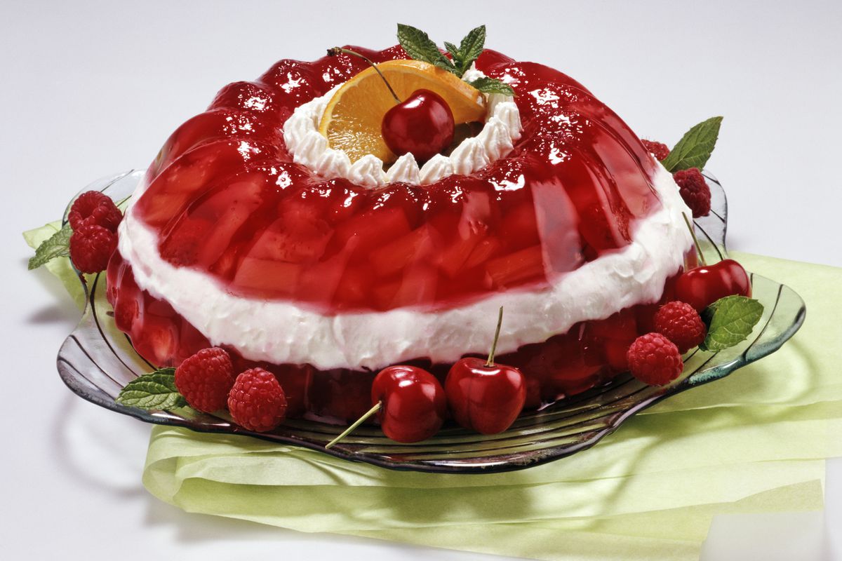 a stock photo showing a molded gelatin dessert with red and white layers on top of a glass dish; it is decorated with cherries around the bottom border and has cream and an orange slice on top