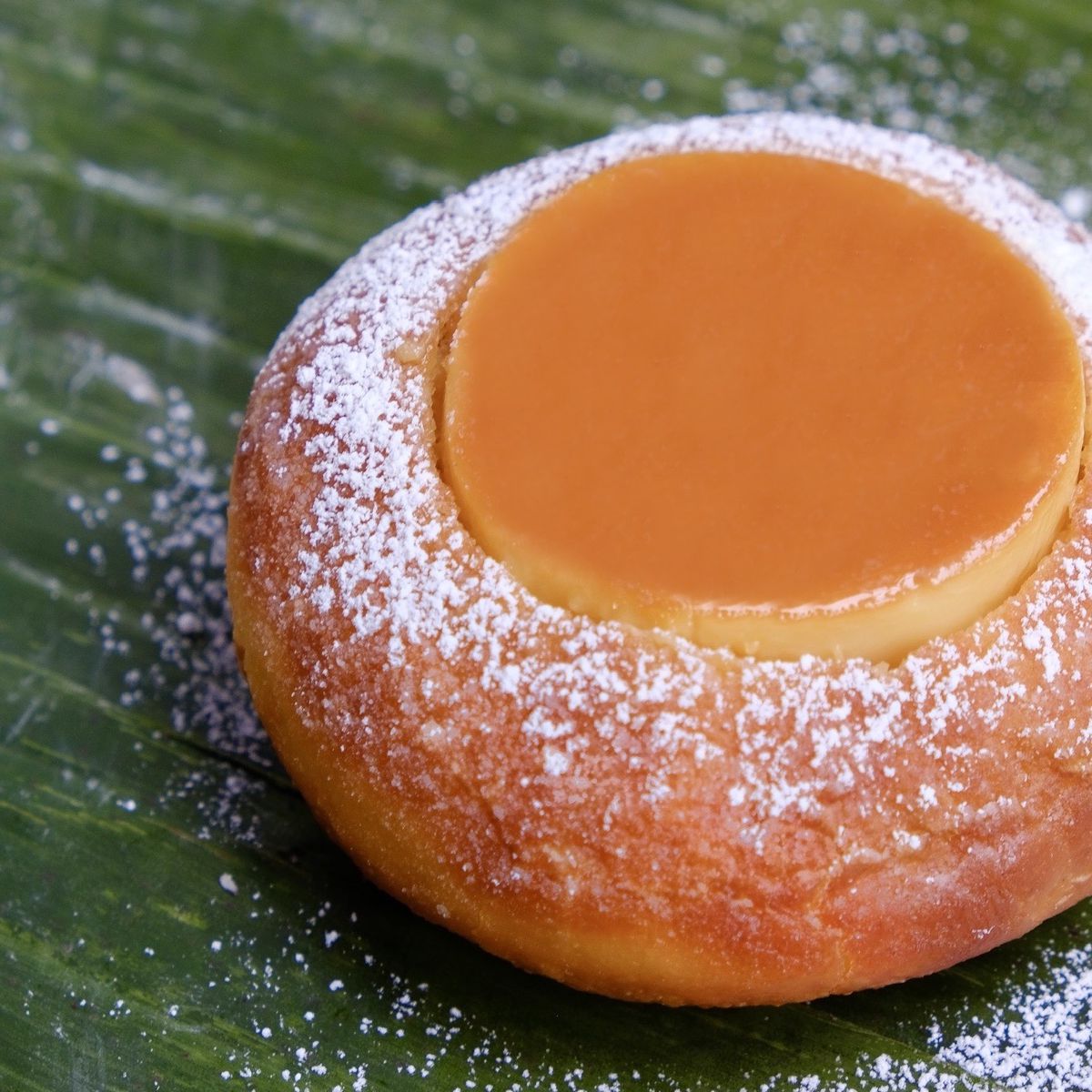 An orange doughnut with a circular flan set in the middle and dusted with white powder on top. The doughnut is set on top of a wide green leaf.