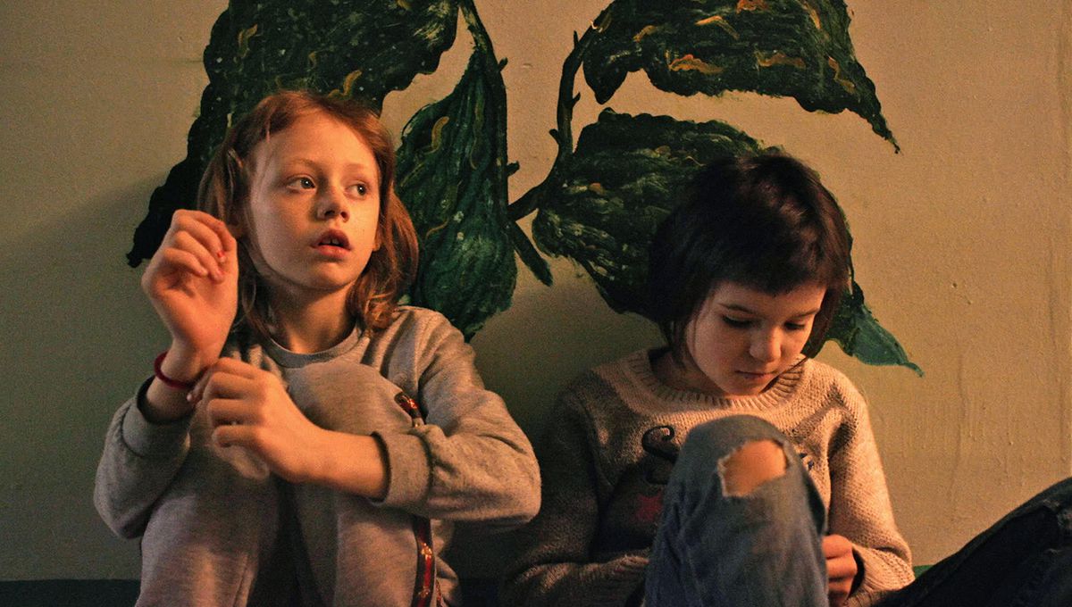 two children sit beside one another against a wall with a green-leaf plant painted on it.