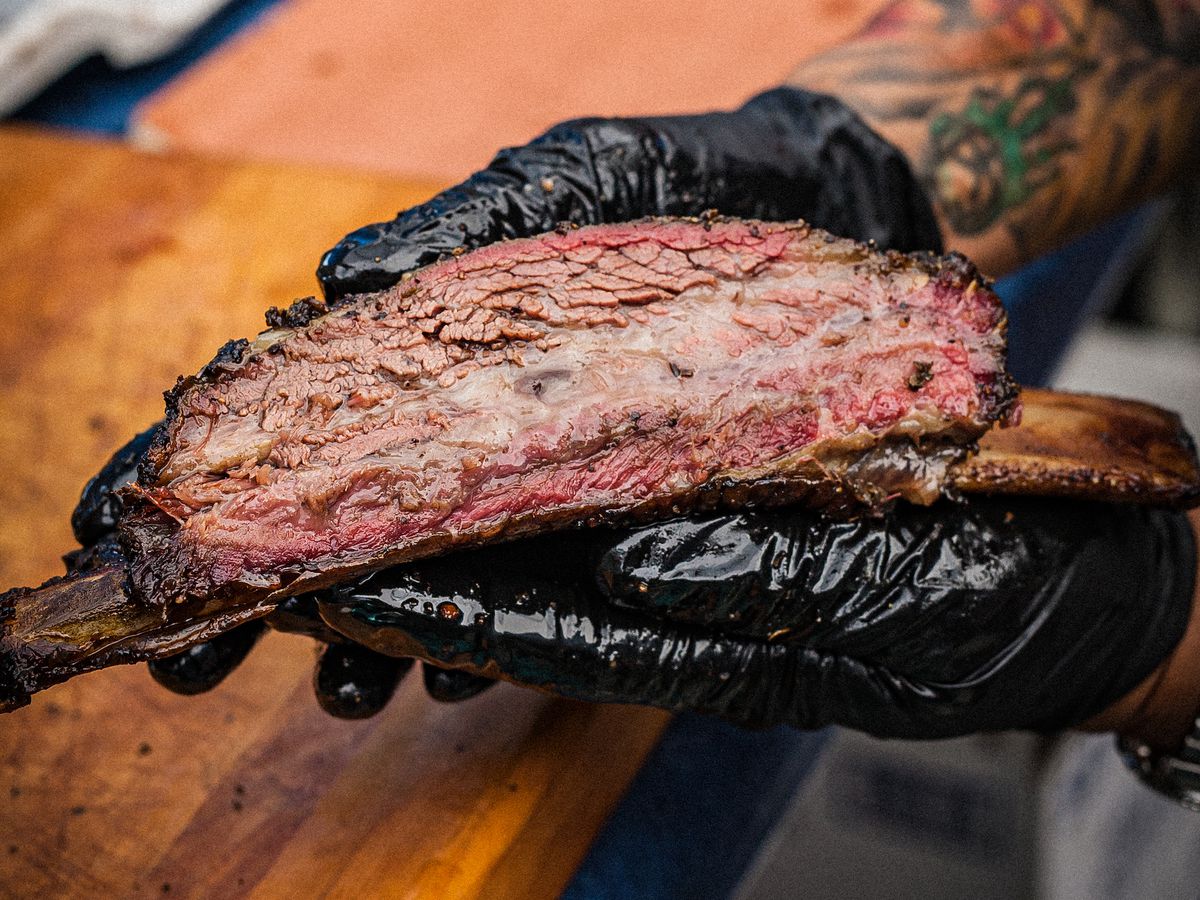A beef rib fresh off the smoker held by black-gloved hands.