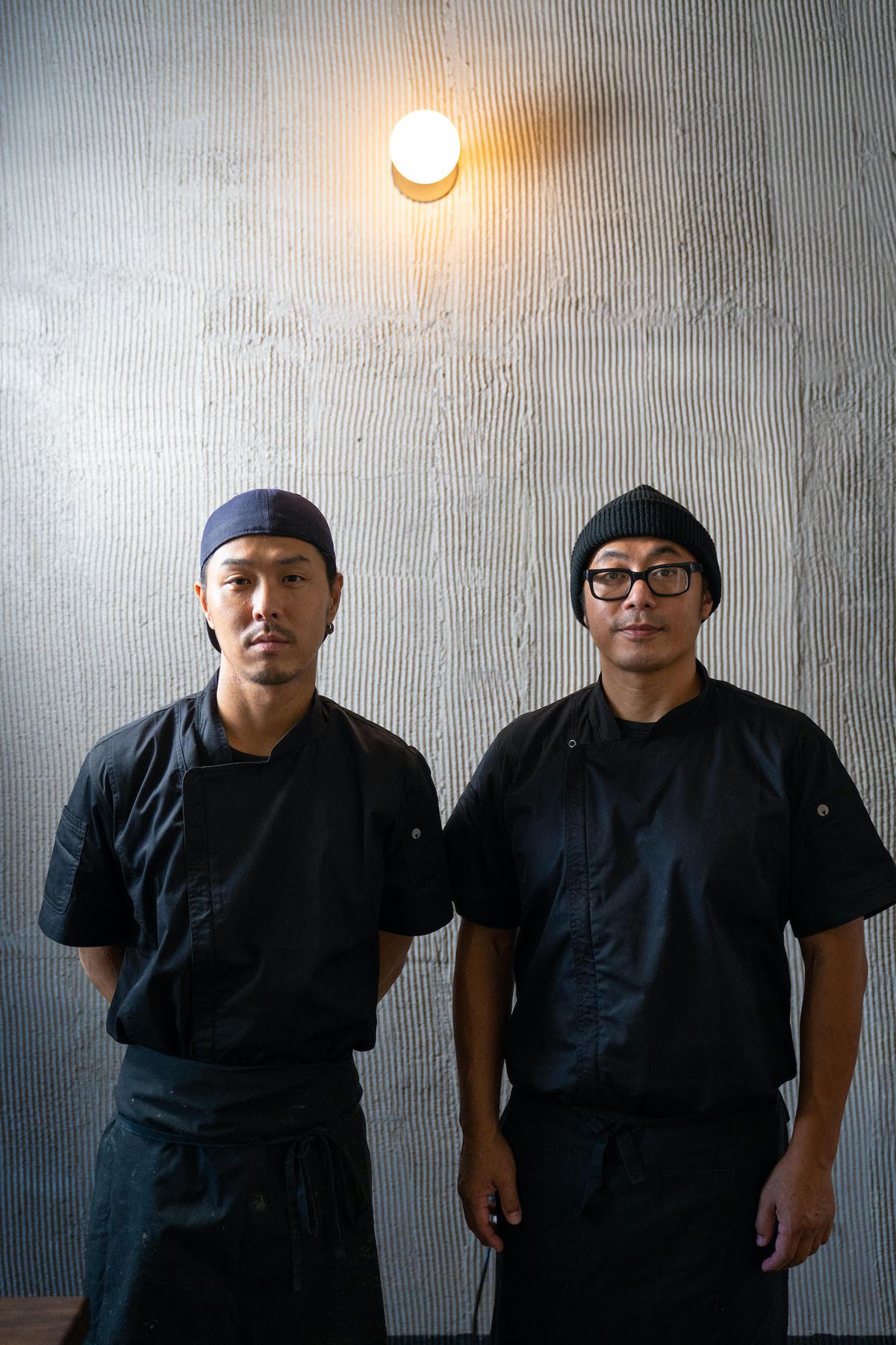 Two Japanese sushi chefs stand inside their small, gray restaurant space.
