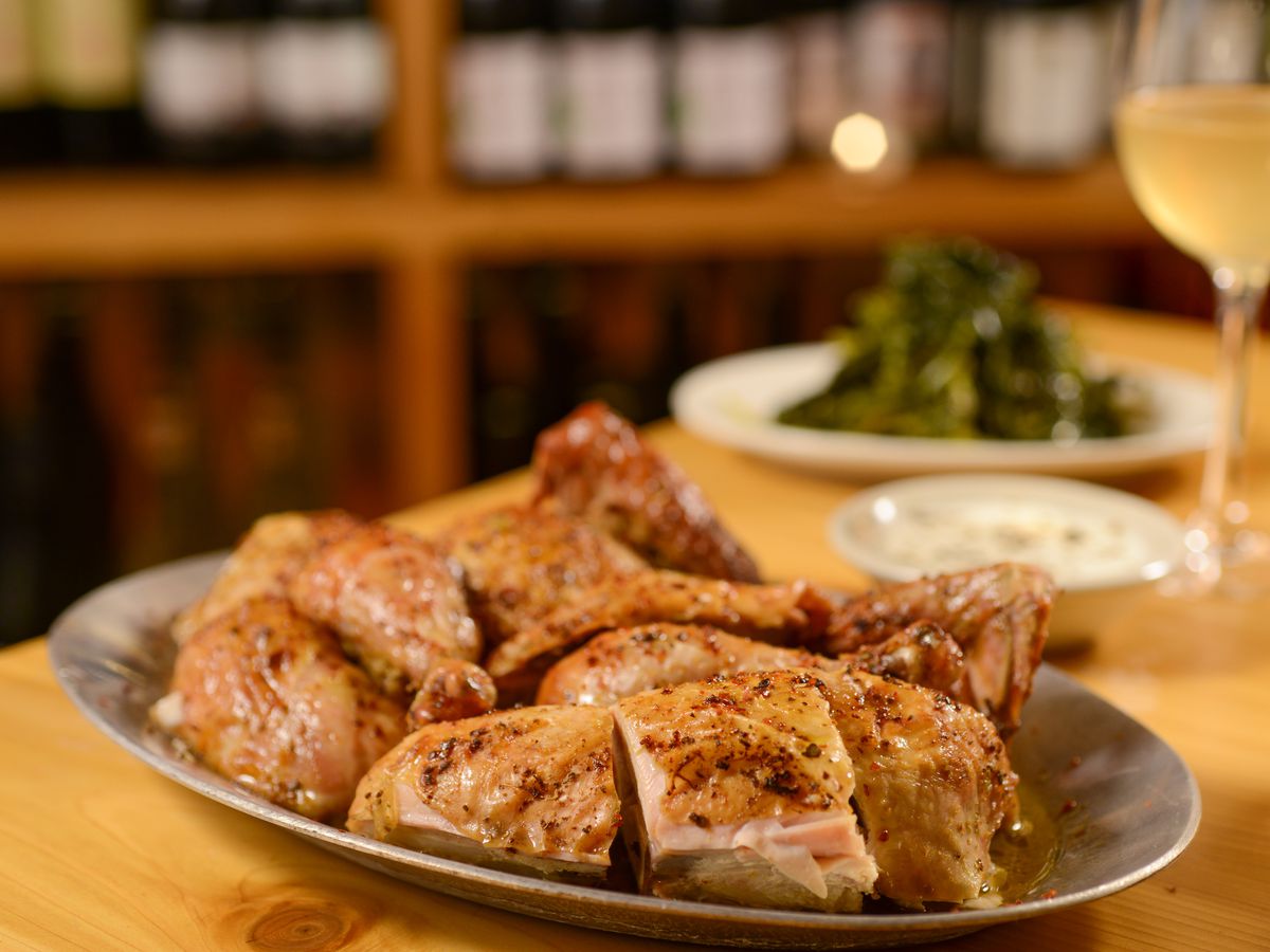 A plate of roast chicken sits on a light wood table next to a glass of white wine.