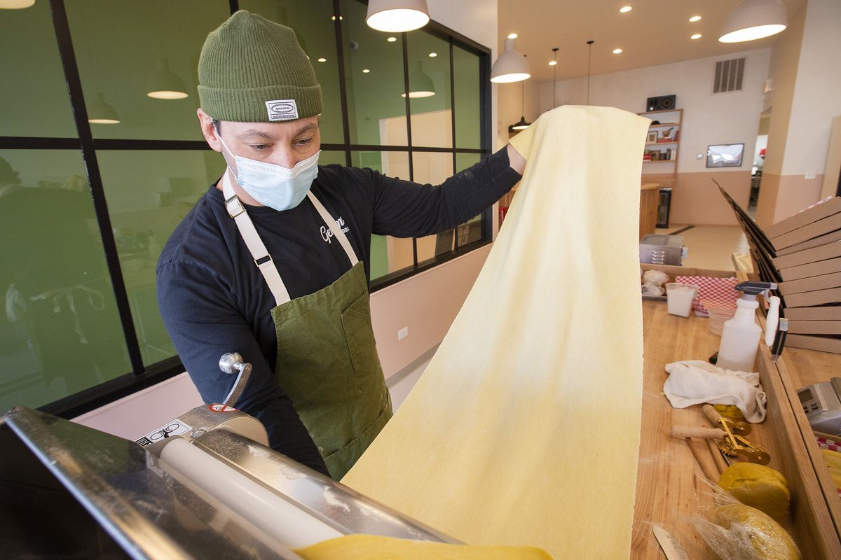 A man in a beanie, a facemask, and an apron removes a sheet of pasta dough from a silver machine.