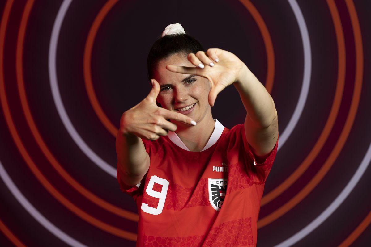 Sarah Zadrazil wears her national team red #9 jersey in a photoshoot, making a camera frame gesture around her face with her fingers.
