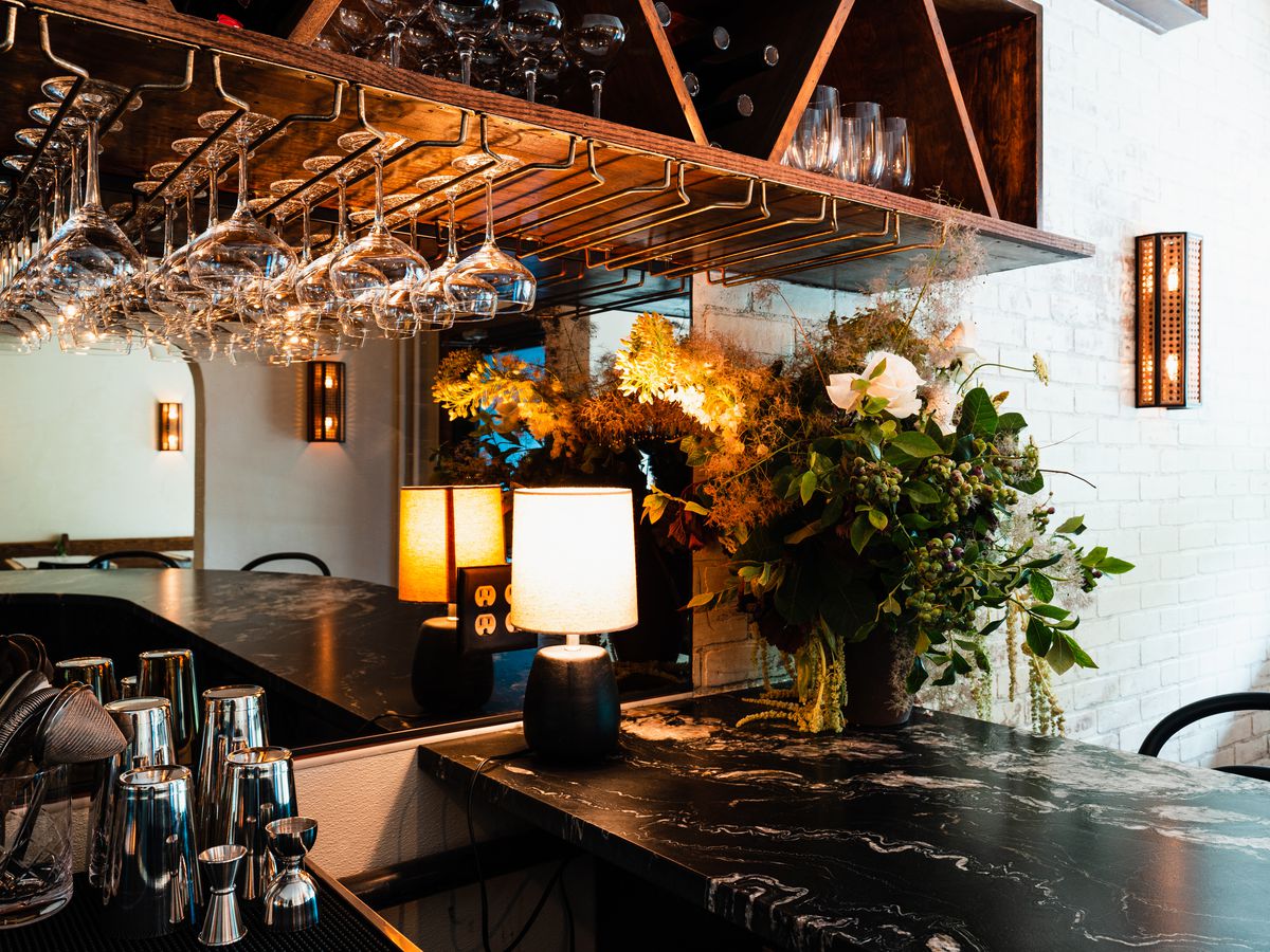 A dark marble bar with a lamp and greenery on it. Above is a wooden rack for wine glasses and wine glasses dangling from it; attached to the wall is a mirror.