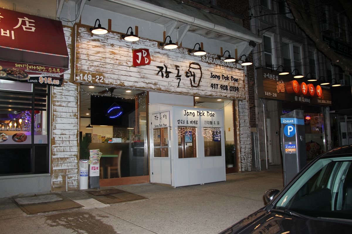 The exterior of a restaurant with a white clapboard front, lights hanging from the entrance, and a tv screen can be seen inside.