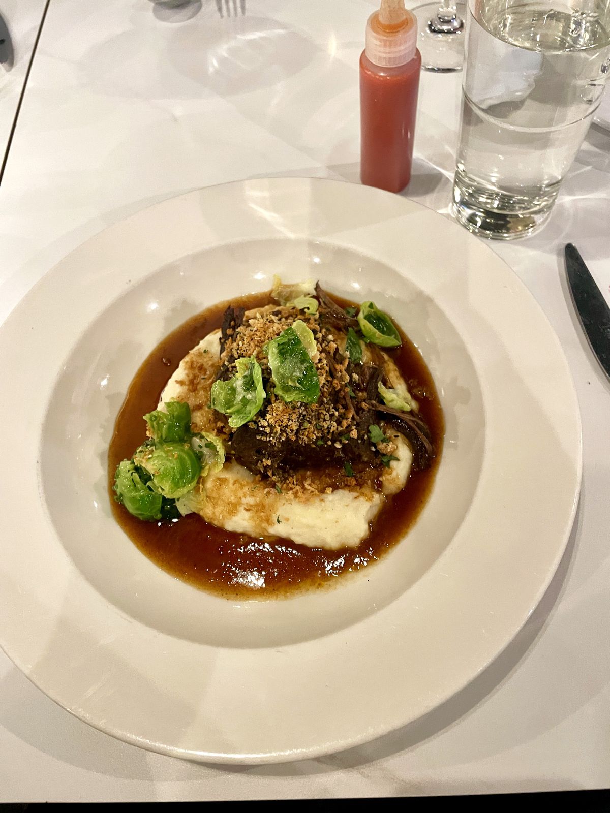 Beef short ribs sit in a bowl atop mashed potatoes. It is sprinkled with Brussels sprouts. A glass of water and a container of hot sauce sit at the top.