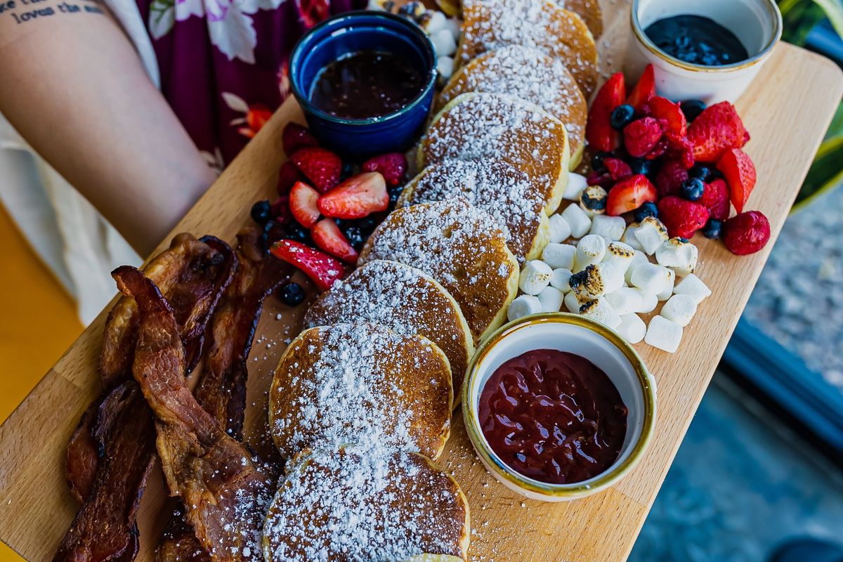 A cutting board with bacon, a bunch of pancakes, strawberries, jams, and syrup.