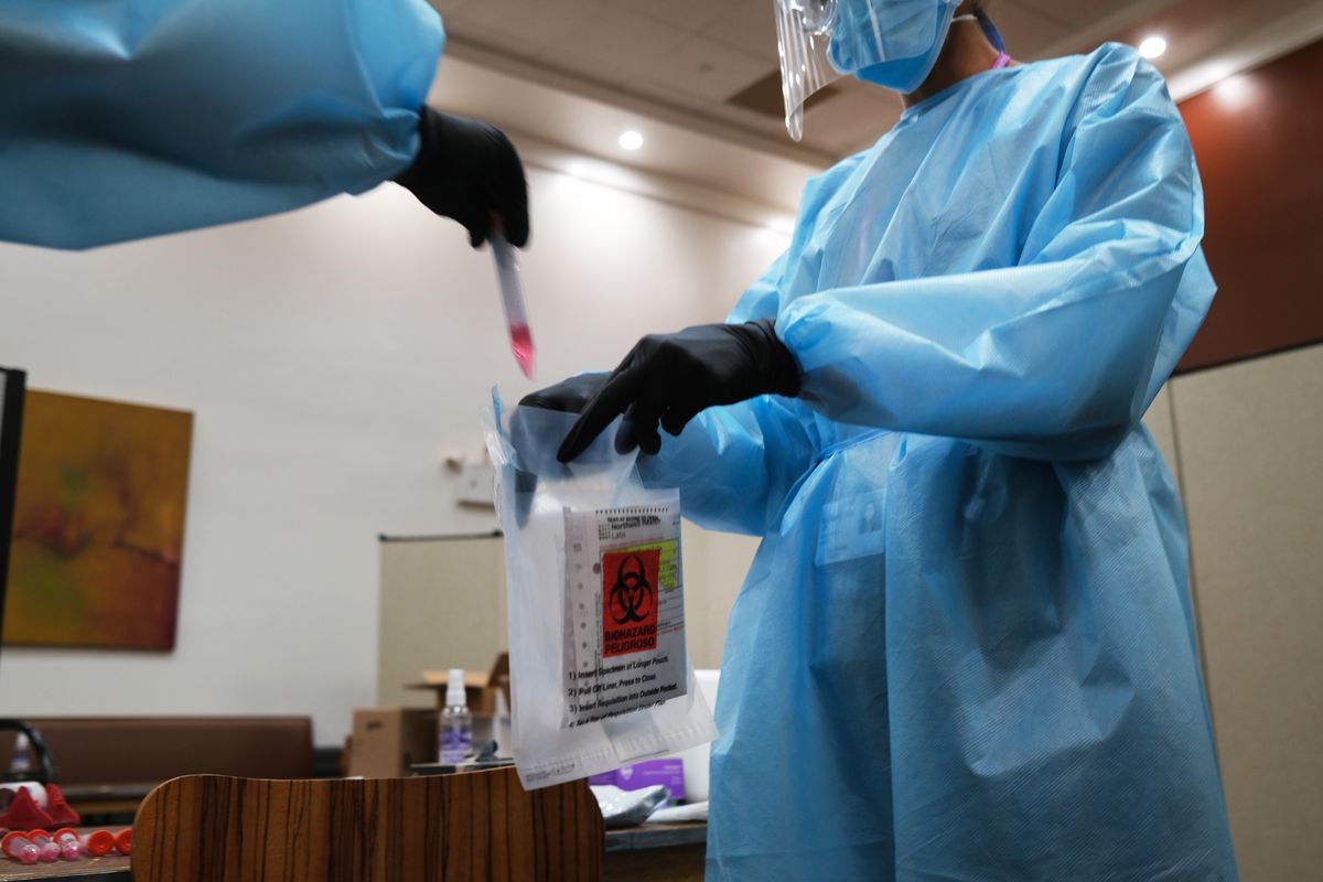 Health workers in gloves, gowns, and face masks bag a testing vial in a biohazard bag.