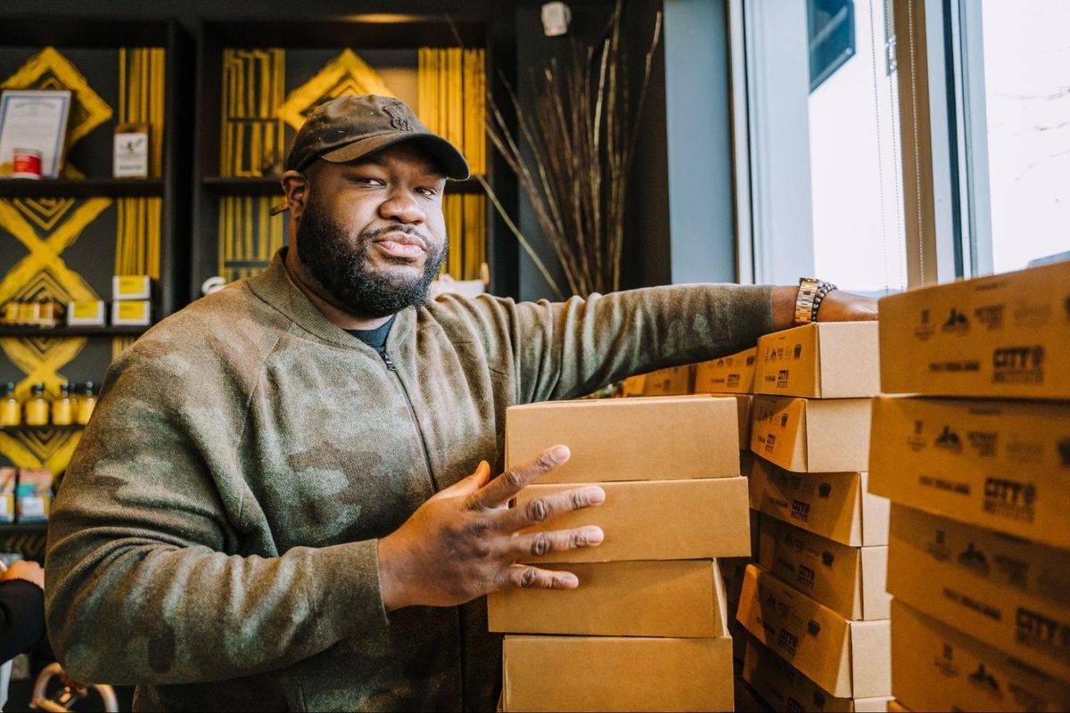 A man staring at the camera, with his hands touching a stack of cardboard boxes filled with meals at Baobab Fare restaurant in Detroit, Michigan.