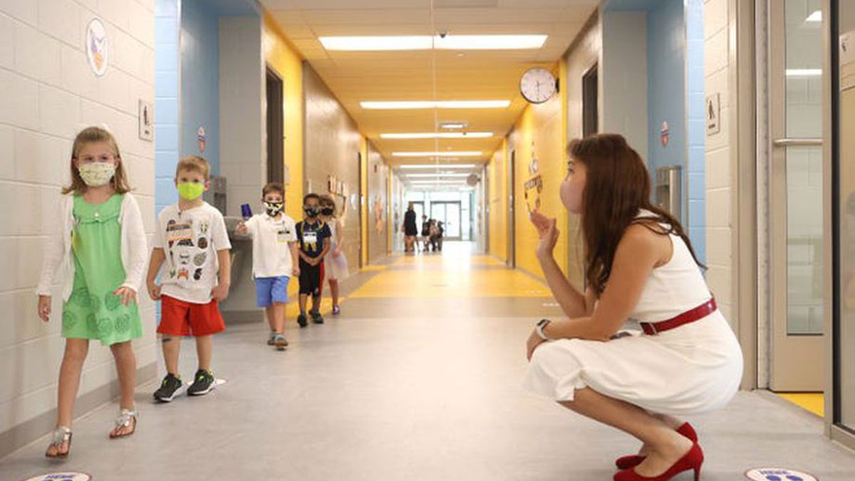 Woman in white sleeveless shift with red patent belt and shoes squats in school hallway, waving at a line of elementary children walking past.