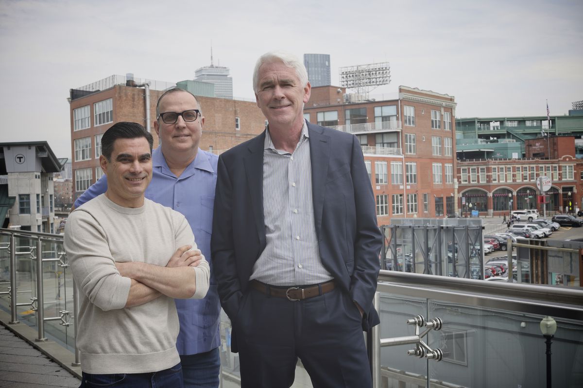 Three men stand smiling outside on a balcony overlooking the Fenway neighborhood. Fenway stadium is visible in the background.