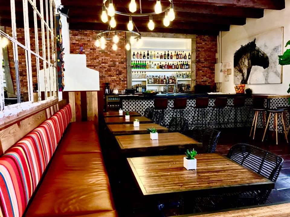 A striped banquette runs alongside one side of a dining room with two tops arranged along it. A large picture of a horse head hangs near a bar toward the back of the room. 