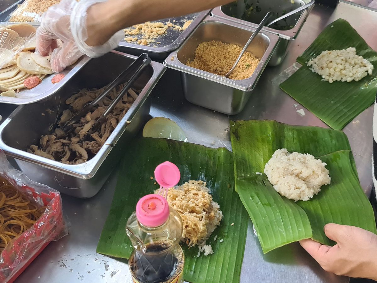 A cook grabs ingredients from prep trays to put together a packet of sticky rice in a banana leaf. 