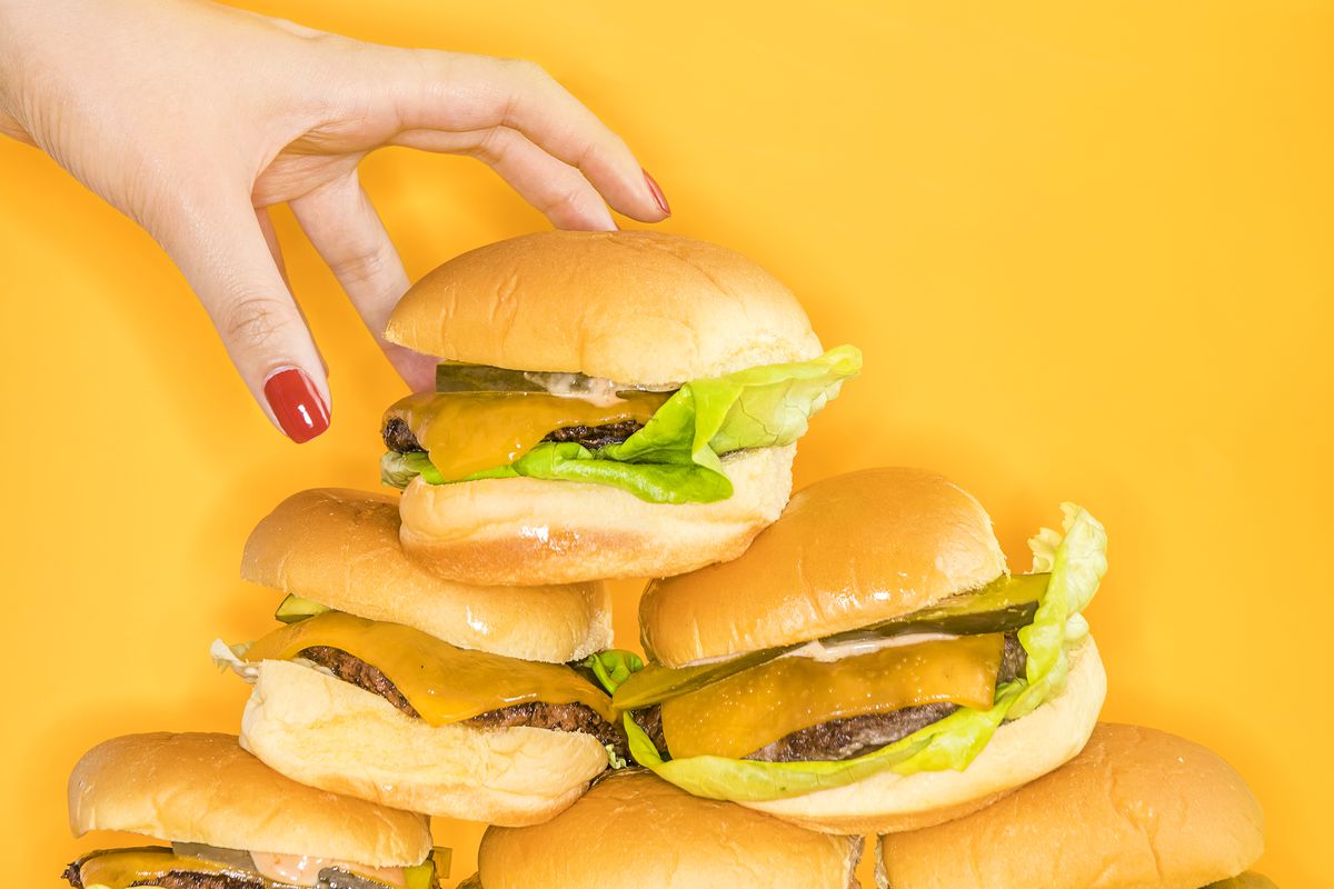 A woman’s red-manicured hand reaches for a pile of cheeseburgers on a yellow background. 