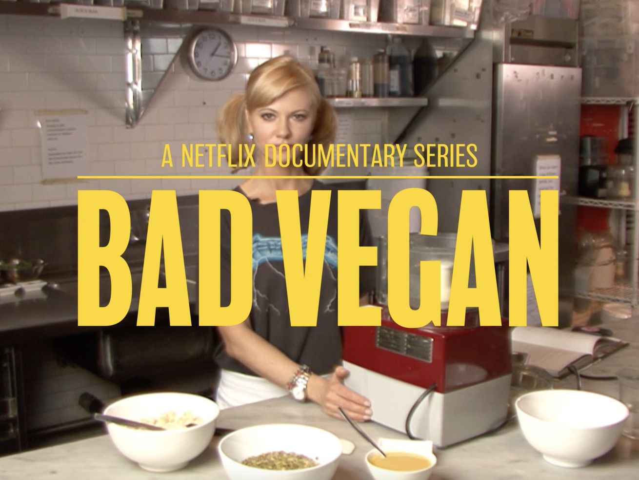 Sarma Melngailis, a thin blonde woman in her 30s, stands in a restaurant kitchen surrounded by white bowls of ingredients and a food processor, with the docu-series title card “BAD VEGAN” in all-caps yellow letters layered over her.