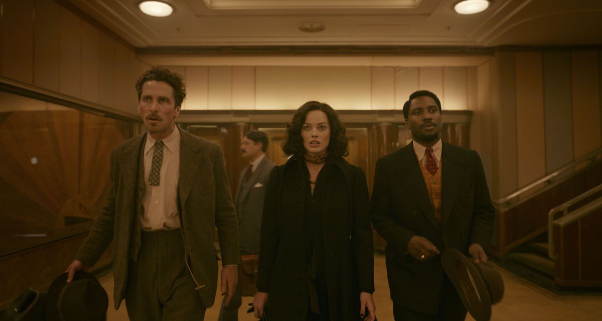 A disheveled and emaciated-looking man (Christian Bale), a dark-haired woman in a black coat (Margot Robbie), and a man in a dark brown suit and tie (John David Washington) walk through a hotel lobby with varnished wood walls.