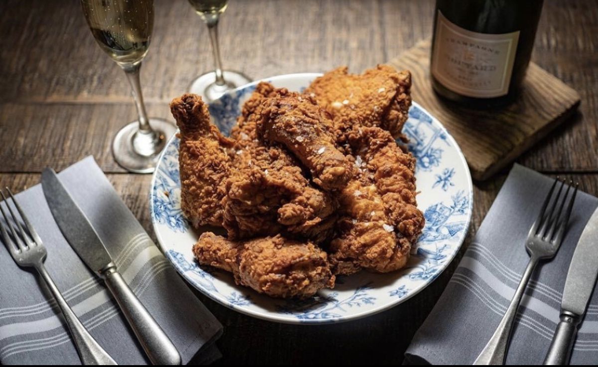 <p style="user-select: auto;">A wooden table set with cloth napkins, Champagne glasses, a bottle of wine, and a toile-patterned plate piled high with fried chicken.
