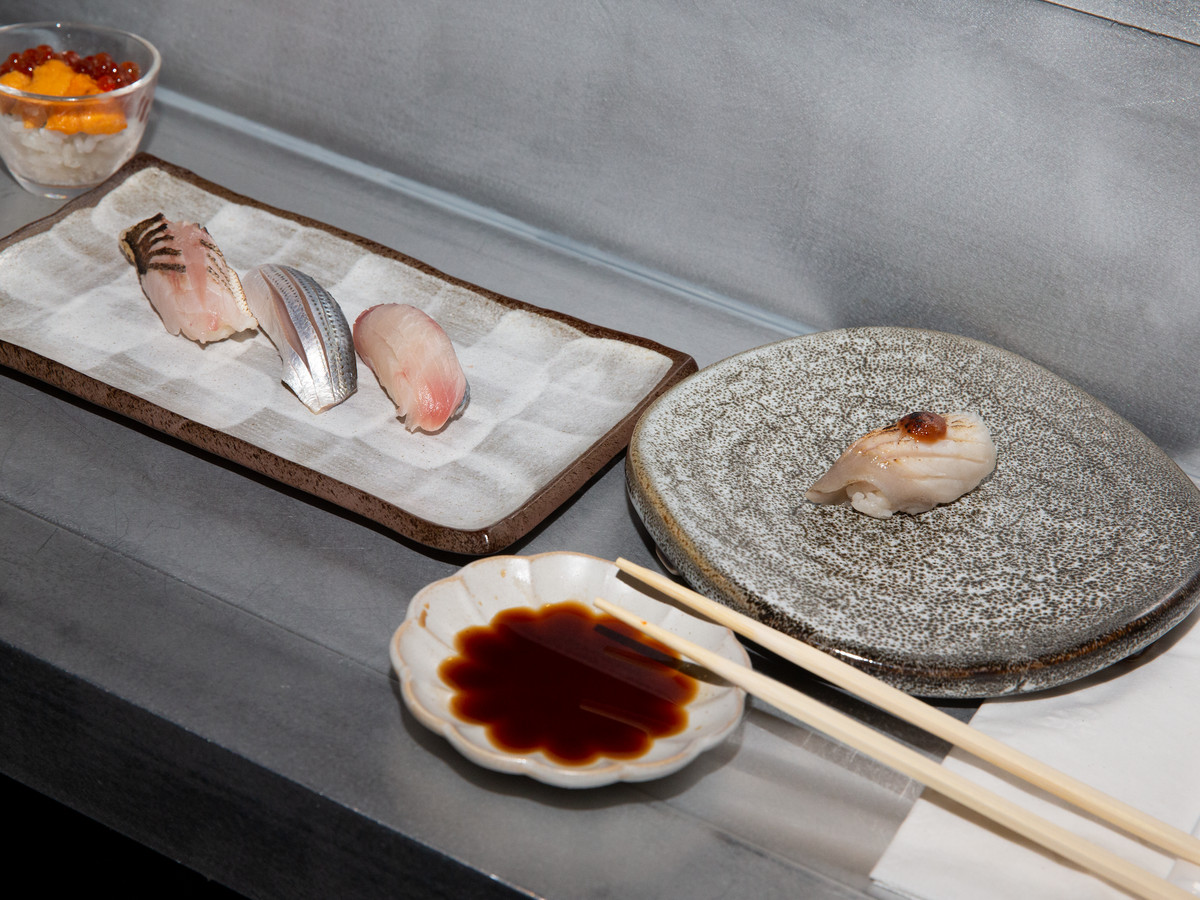 A rectangular plate with three pieces of sashimi, a round plate with one piece of sashimi, and a small bowl with soy sauce and resting chopsticks all on a gray counter.
