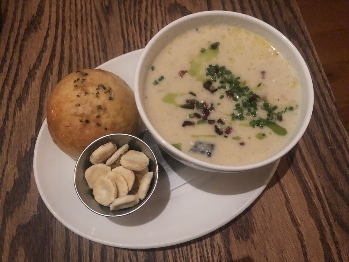 A white dish filled with soup with a cup of crackers and a dinner roll placed beside it on a white plate.