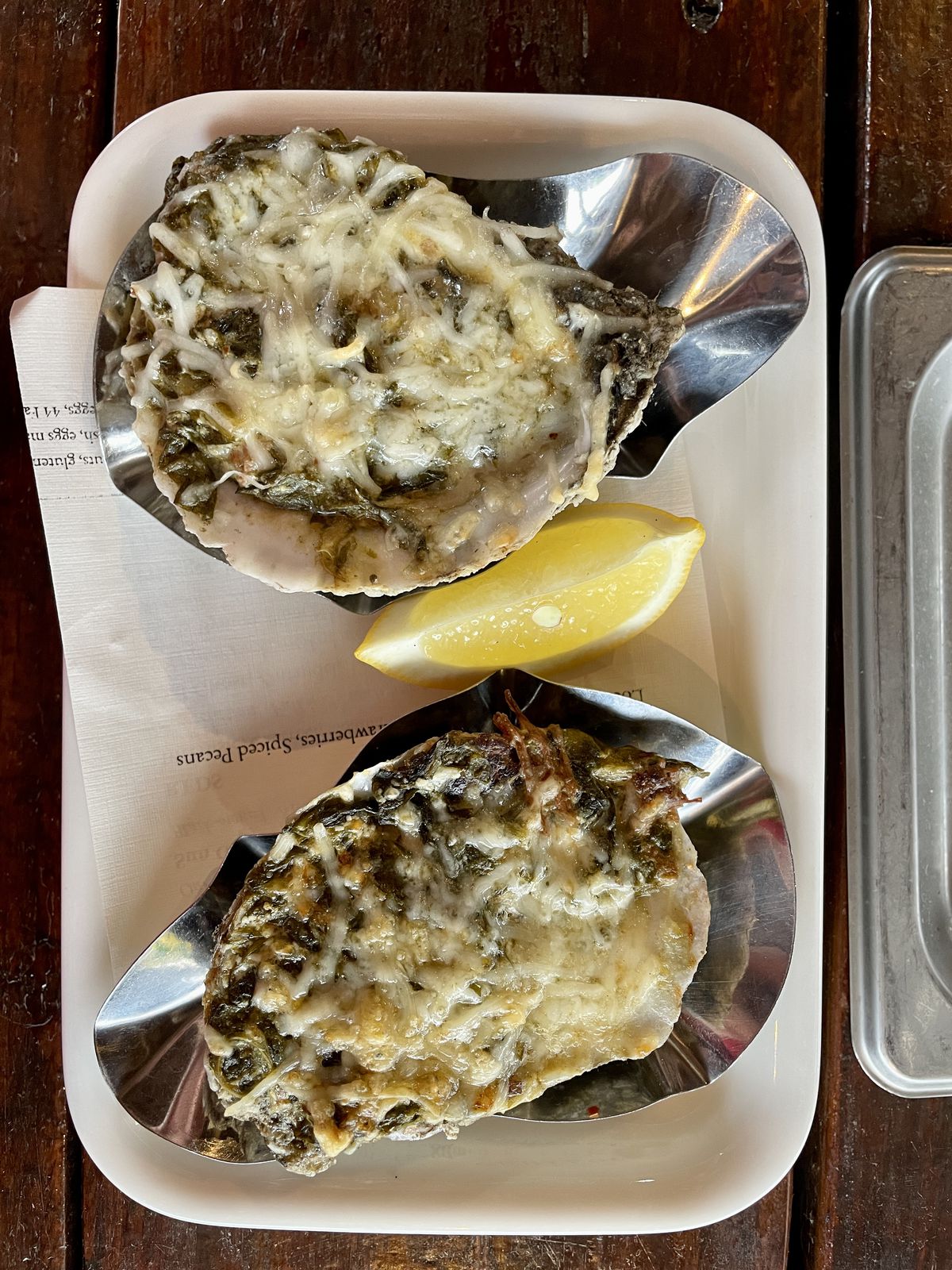 A white dish holds two tins with large baked oysters covered in parmesan cheese.