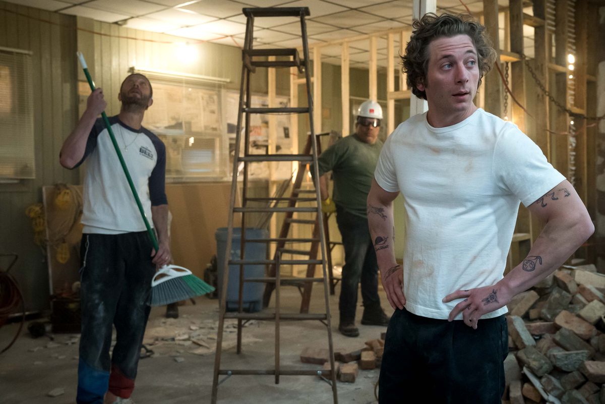 Carmy stands in the foreground of his gutted restaurant as Richie holds a broom and looks to the ceiling and a man in a hard hat stands behind a stepladder in season 2 of The Bear