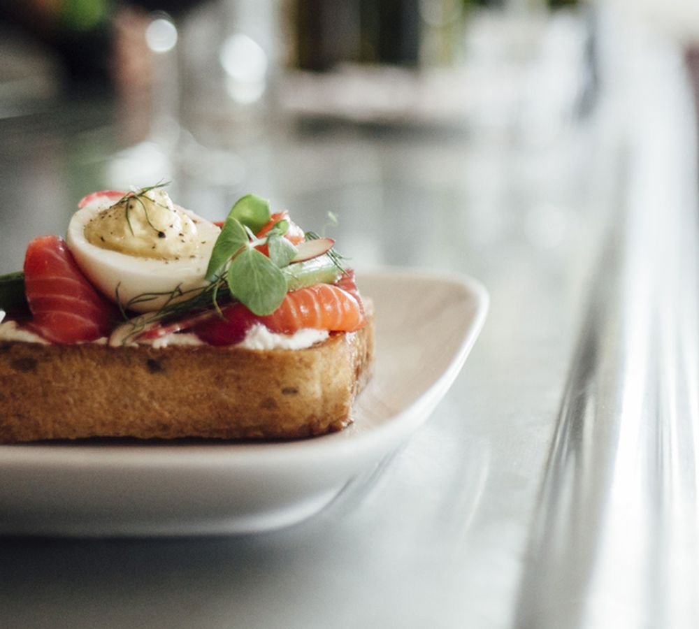 A piece of toast topped with cream cheese, lox, herbs, and an egg on a plate that’s sitting on a long counter. 