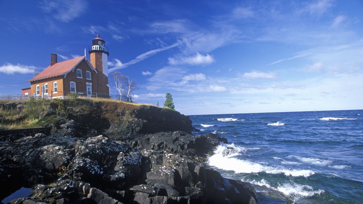 A red brick building attached to a lighthouse overlooking a rocky cliff bashed by waves