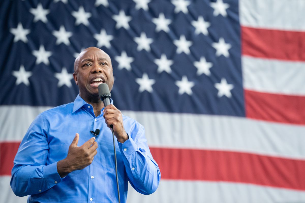 Sen. Scott, a Black man wearing a blue Oxford shirt, speaks into a handheld microphone in front of a large American flag backdrop.