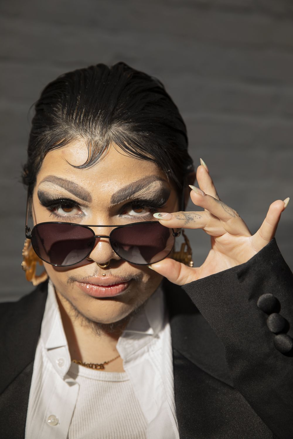 Closeup of a performer with sparse mustache and beard and long nails lowering aviator sunglasses and glancing at the viewer