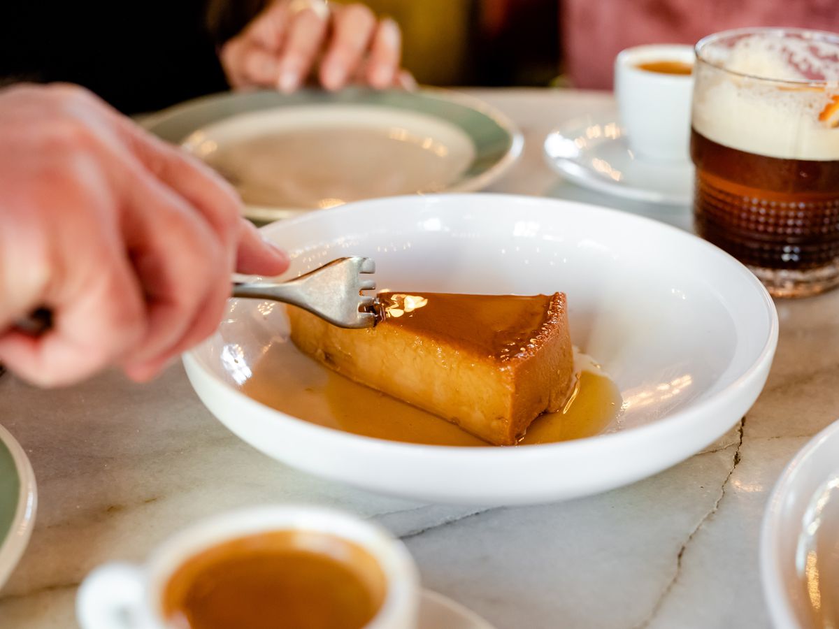 A hand holding a fork that’s slicing into a piece of flan.