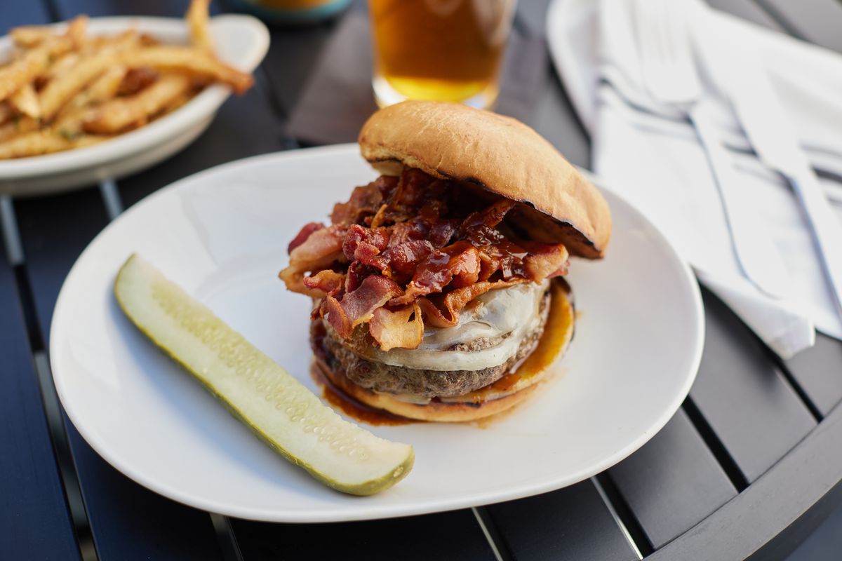 A burger topped with bacon and white cheddar sits on a white plate beside a dill pickle spear. In the background are french fries and a glass of beer. 