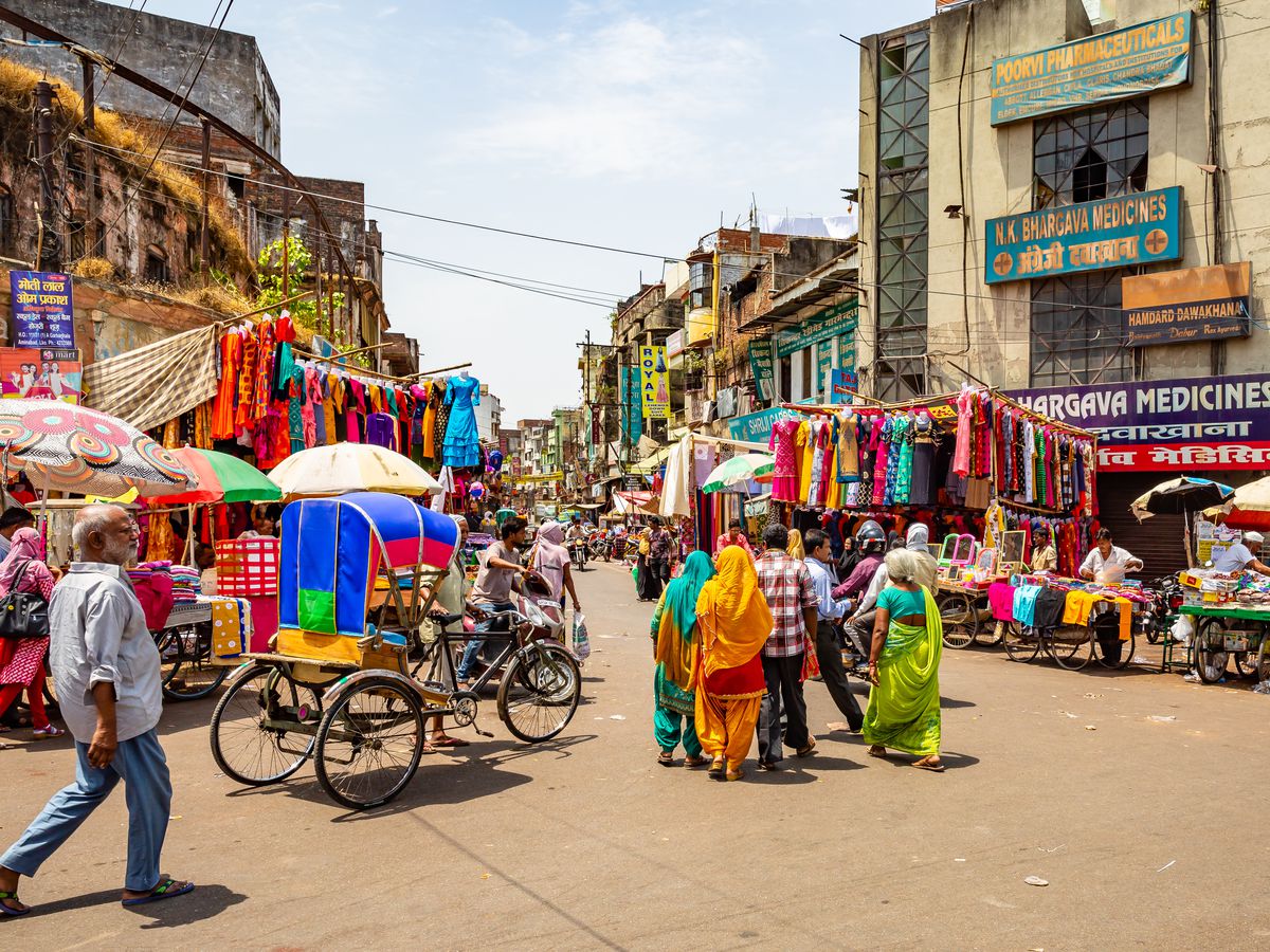A market takes over a street in Delhi, with vendors selling colorful clothing receding into the distance in the center, an empty rickshaw to one side, and groups of locals walking.