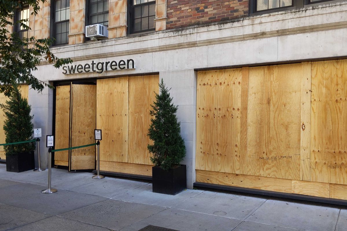 The ground floor of a building with plywood covering all the windows and a sign reading “Sweetgreen” out front