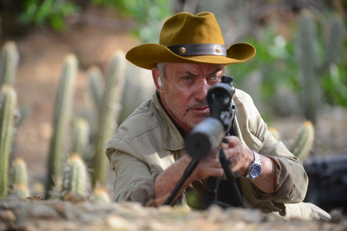 Udo Kier, in hunting khakis and a mustard-yellow bush hat, lies in a desert, surrounded by cacti, and pointing a braced sniper rifle into the camera.