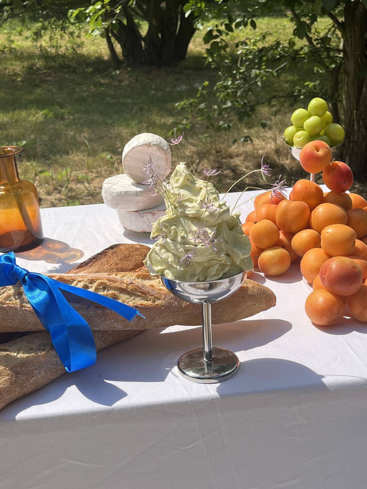 on a white table outdoors is an artistic spread by the culinary studio Herrlich Dining. a metal coupe glass of softened butter with flowers poking out sits front and center, with a pile of peaches to its right and two baguettes tied with a blue ribbon to its left. in the background are stacked wheels of cheese