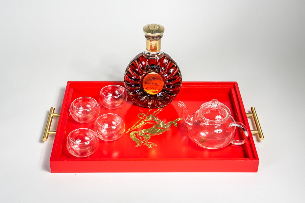 A red tray with gold handles holds four glass teacups, a glass teapot, and a bottle of Rémy Martin.