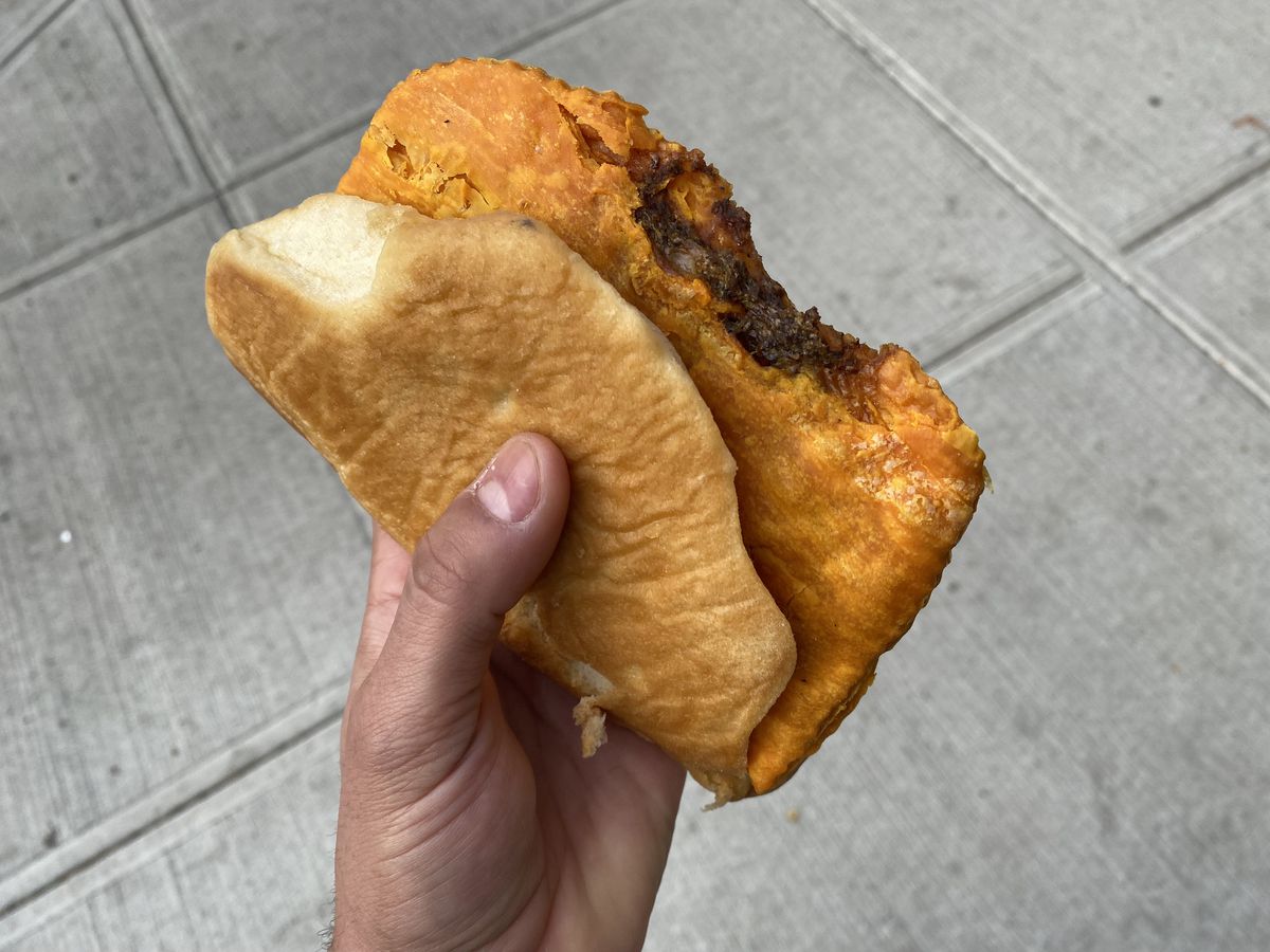 A hand clutches an orange beef patty with cheese tucked into a piece of coco bread.