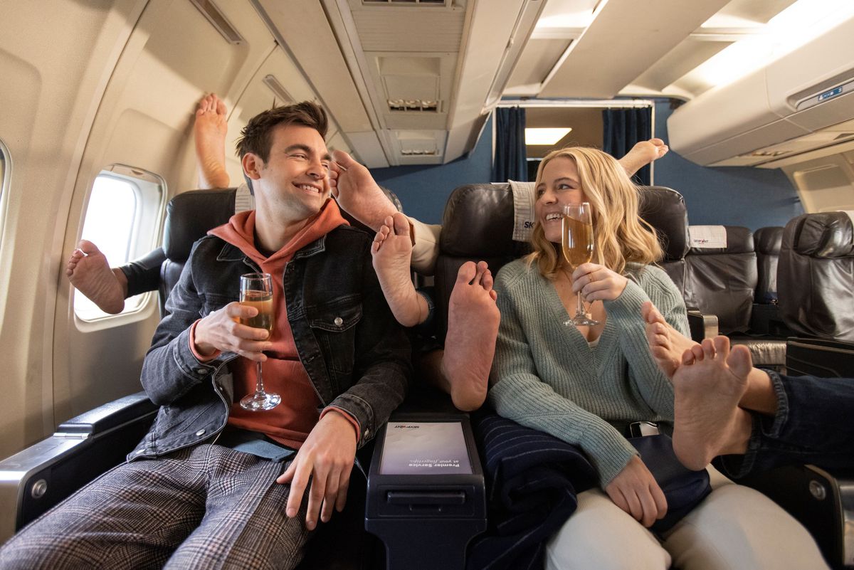 Two people sitting in an airplane seat with feet filling in the gaps around them in a still from season 2 of The Other Two